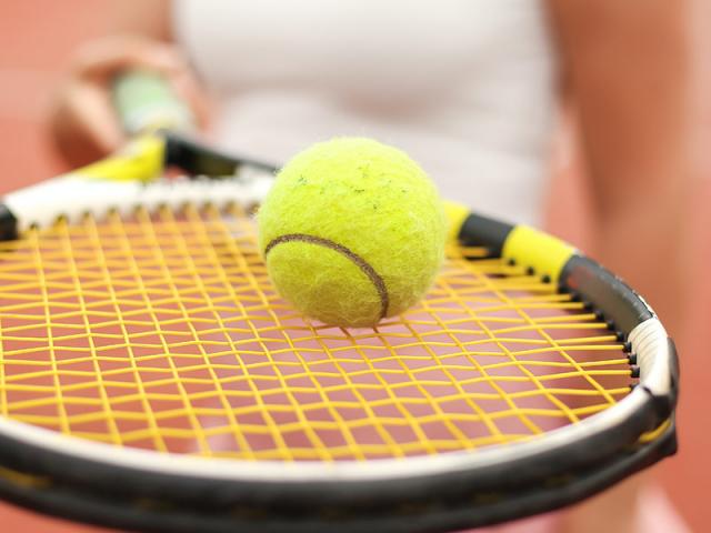 A closeup photo of a tennis ball, balanced on a racket, held by a woman in the background