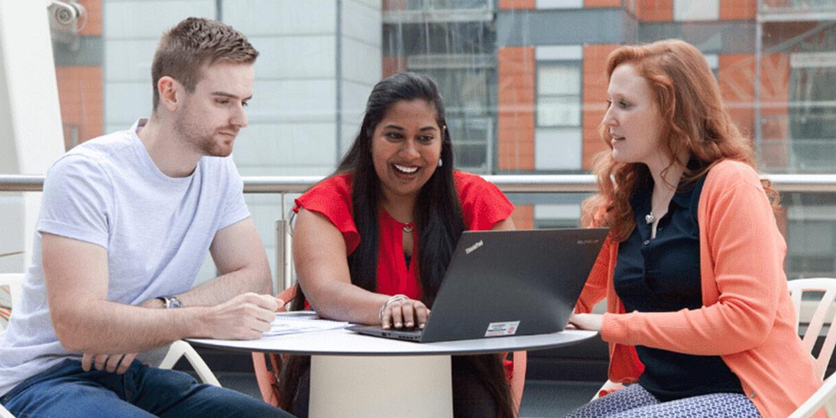 A man and two women sit around a table looking at a laptop