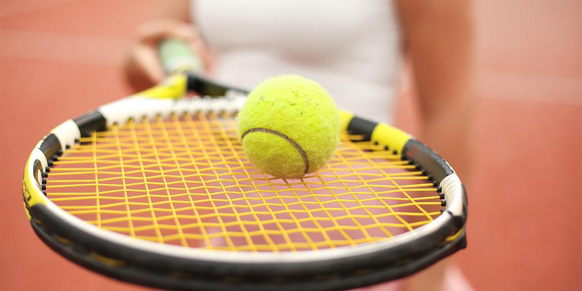 A closeup photo of a tennis ball, balanced on a racket, held by a woman in the background