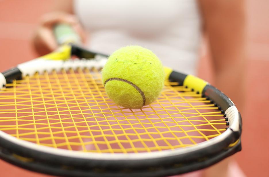 A closeup photo of a tennis ball, balanced on a racket, held by a woman in the background